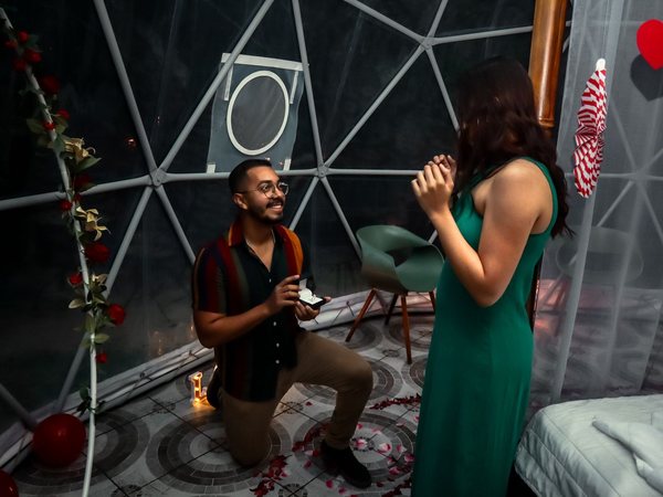 Hombre arrodillado proponiendo matrimonio con anillo dentro del domo transparente decorado con petalos y velas.