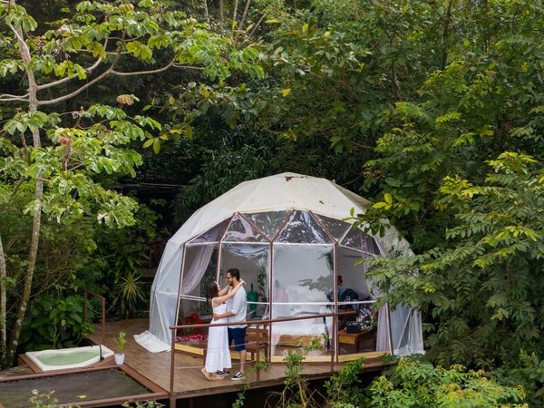 Pareja abrazada en el deck del domo panoramico con jacuzzi rodeado de selva.